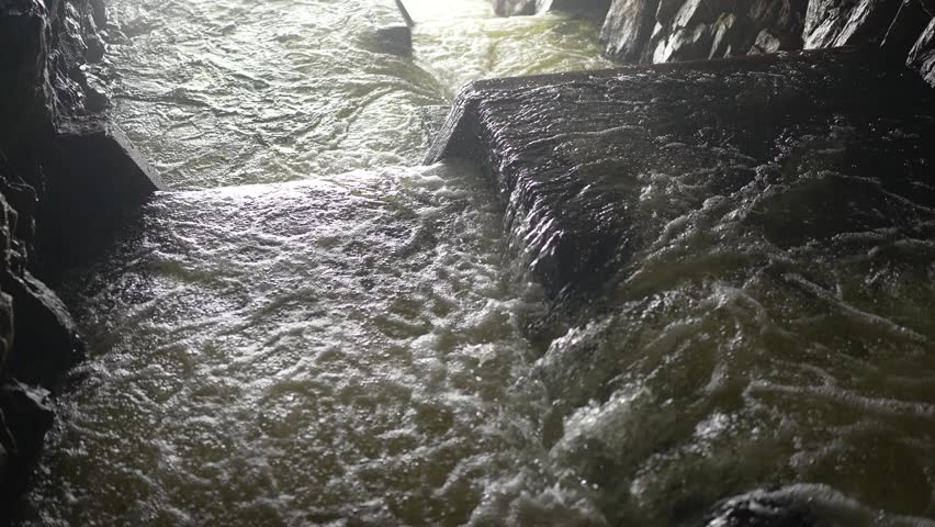 Water rushes rapidly through a rocky channel, creating a turbulent flow as it moves in the dimly lit underground space. The sound of rushing water echoes in the air.