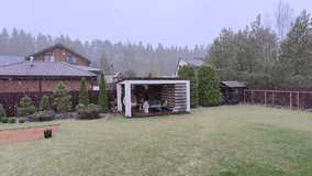A wooden gazebo with a table, benches and swings stands next to a grassy lawn. Behind it are a fence, trees, buildings and forest. It is snowing heavily in the middle of spring. Overcast weather - Powered by Shutterstock - Get 15% off with code: PIKWIZARD15