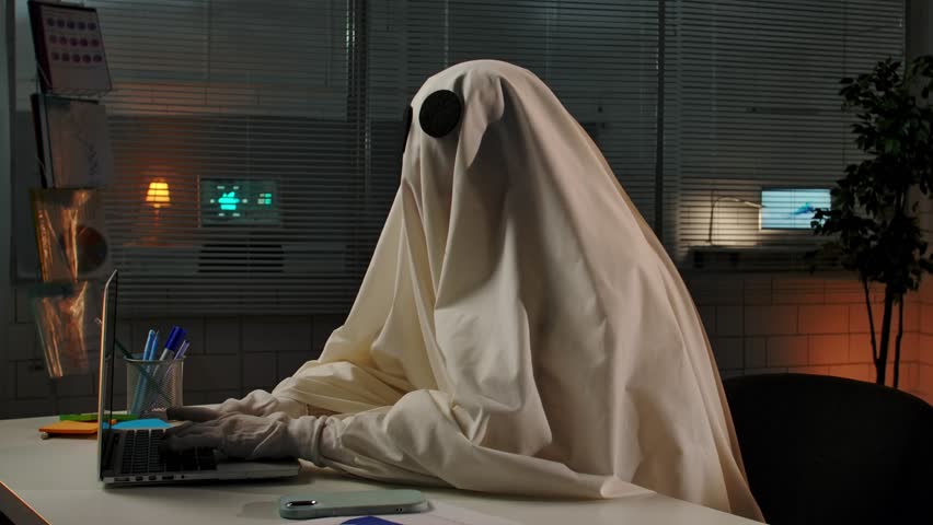 A person dressed as a ghost in an office with a laptop and a phone, creating a humorous and spooky atmosphere.