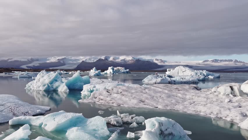 Aerial shot showcases icebergs of various sizes and shades of blue and white, densely packed within the vast Jökulsárlón glacier lagoon, with a backdrop of rugged, snow-capped mountains.
