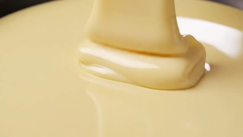Close-up of creamy white chocolate pouring smoothly, forming soft folds and glossy ripples in a rich beige-yellow tone.