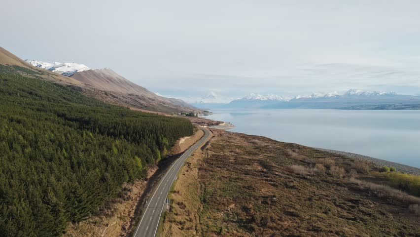 A slow aerial push-in over State Highway 80 in New Zealand, leading toward snow-capped Mount Cook with Lake Pukaki stretching alongside under a clear morning sky
