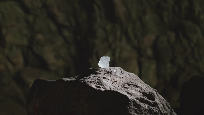 Cinematic shot of a luminous raw crystal resting on a rock pedestal in a dark cave. Mysterious smoke drifts by, creating a magical or sacred fantasy atmosphere.