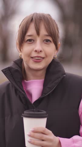 Cheerful young woman sipping steaming takeaway coffee from disposable cup experiencing a moment of pure enjoyment on a cool day outdoors, warm and looking at the camera vertical video