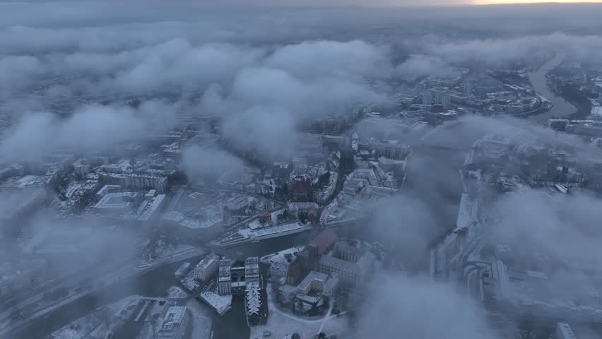 An aerial winter view of Wroclaw, Poland, with snowy rooftops and thick clouds partially veiling the city