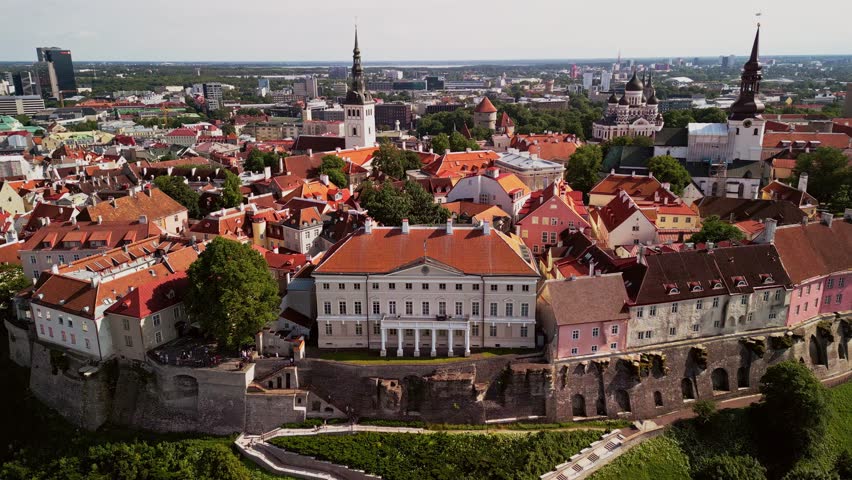 Aerial drone footage captures the historic charm of Tallinn Old Town and Toompea Hill under a bright sunny sky, showcasing ancient red rooftops, cobbled streets, and medieval towers.