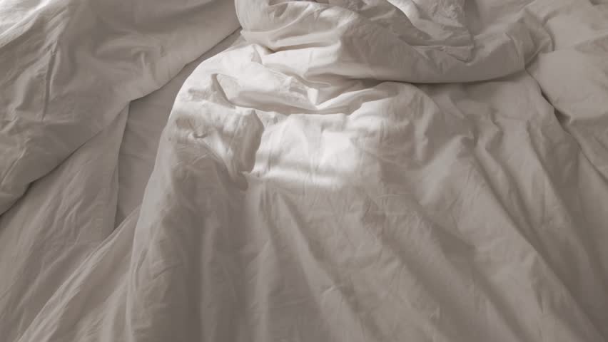 Soft morning sunlight casting shadows on wrinkled white cotton bedsheets. Morning vibes, unmade empty bed. Minimal and cozy home interior. Messy bedroom in a hotel or in an apartment.