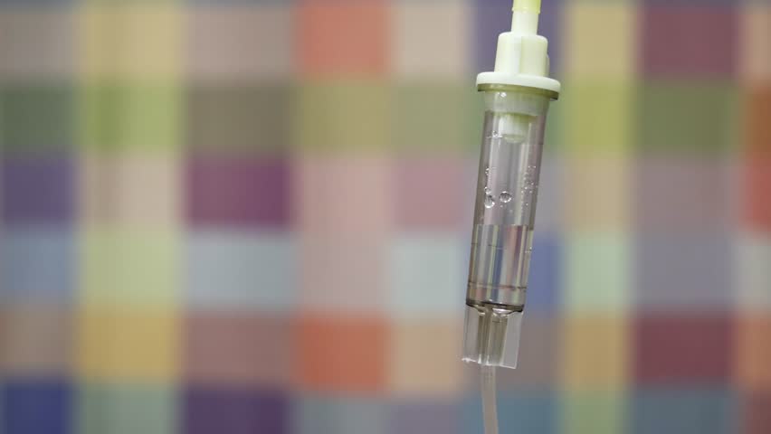 A closeup shot of an IV drip chamber with visible fluid drops, set against a blurred multicolored curtain - Powered by Shutterstock - Get 15% off with code: PIKWIZARD15