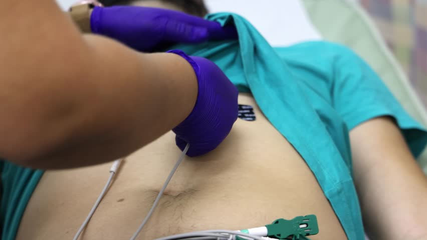 A healthcare worker performs an ECG on a patient's chest using electrodes while wearing purple gloves