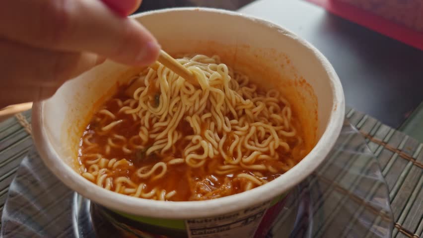 person using chopsticks to lift steaming noodles from cup of instant ramen, showcasing a quick and comforting meal. rich broth and curly noodles Asian fast food. Instant noodles cup. Budget food
