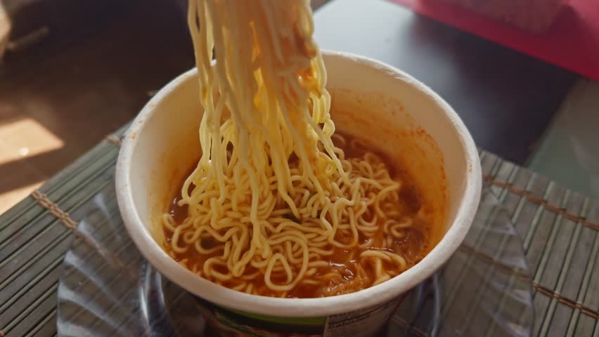 person using chopsticks to lift steaming noodles from cup of instant ramen, showcasing a quick and comforting meal. rich broth and curly noodles Asian fast food. Instant noodles cup. Budget food