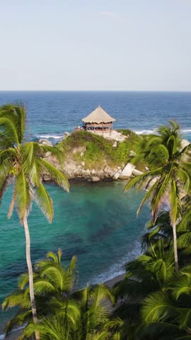 Aerial view of Cabo San Juan del Guia in Tayrona National Park, Colombia, featuring a thatched roof kiosk on a small hill surrounded by palm trees and turquoise water