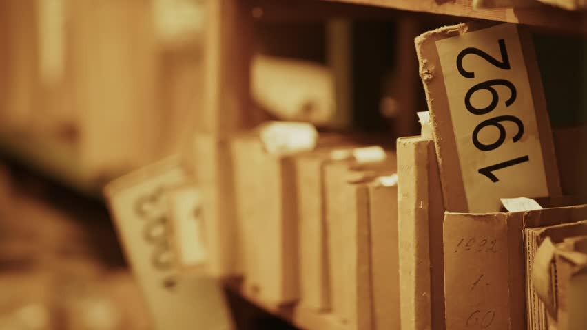 Old archive room with shelves filled paper files folders organized by year. Worn documents suggest historical or governmental records. Research files document storage old records. Archival stacks file