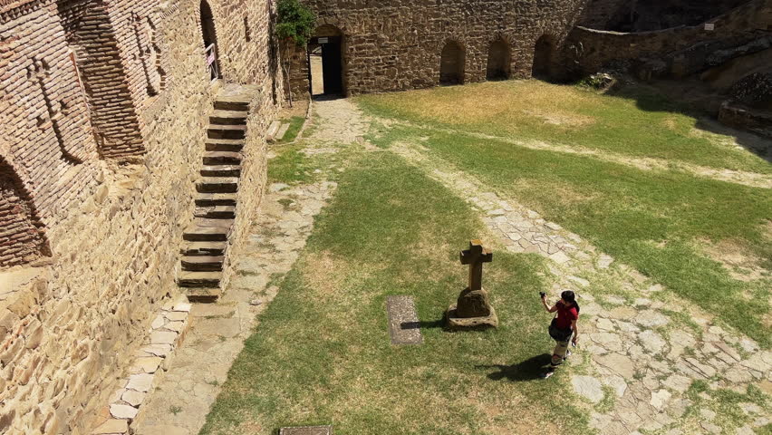 Downward shot captures traveler snapping photo at cross monument, with weathered stone walls and golden grasslands stretching across. Ideal for pilgrimage or heritage content.