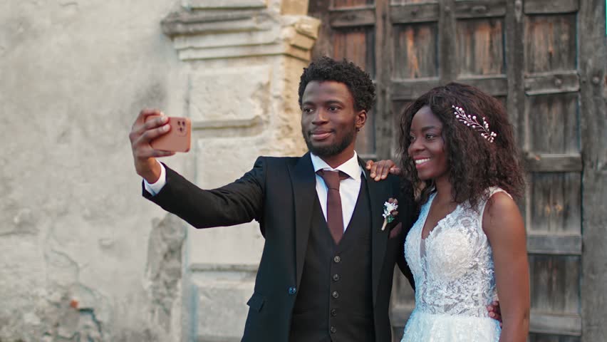 Groom winking and taking selfie on phone with smiling bride in wedding dress near rustic wall. African American couple enjoying wedding moment, capturing memories with smartphone in fun outdoor photo.