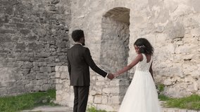 Bride and groom holding hands while walking, groom glancing back smiling. African American couple enjoying romantic moment near ancient stone wall during wedding photo session outdoors. - Powered by Shutterstock - Get 15% off with code: PIKWIZARD15