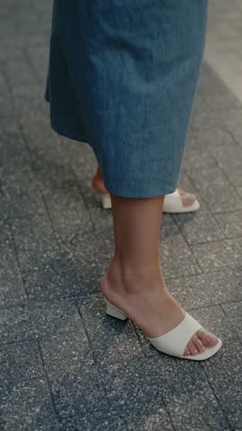 Close-up of women's feet in stylish white open-toe heeled sandals. They stand on a gray paved walkway. The elegant shoes and casual, flowing skirt create a chic summer look.