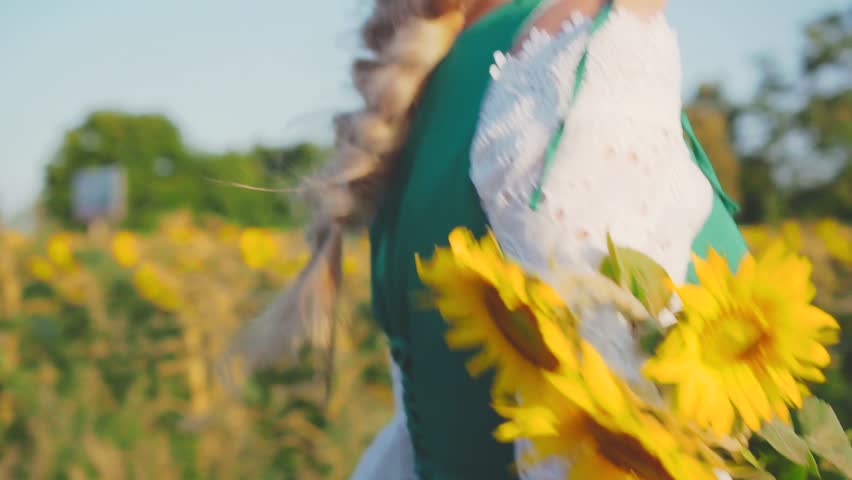 happy young beautiful blonde woman holds sunflower yellow flower in hands walking along dirt country road. summer blooming field sunflowers. joyful girl princess dress vintage style back rear view.