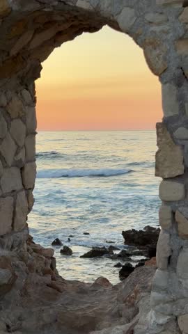 Golden sunset seen through a stone arch in the Old Town of Chania, Crete. Waves gently crash against the rocky shore as warm light fills the historic coastal ruins.