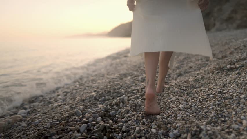 Barefoot woman in white dress walking along pebble beach during golden sunset, embodying peaceful travel and serene coastal exploration