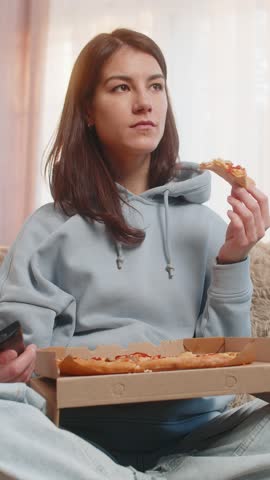 Happy young Caucasian woman eating delicious junk food pizza and popcorn sits on couch sofa watching TV show or movie in home living room. Smiling girl in casual clothes enjoying weekend in apartment