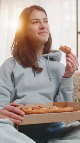 Happy young Caucasian woman eating delicious junk food pizza and popcorn sits on couch sofa watching TV show or movie in home living room. Smiling girl in casual clothes enjoying weekend in apartment