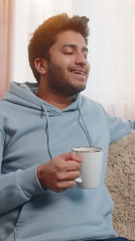 Smiling Indian man drinking warm coffee or herbal tea at home in living room. Hispanic guy enjoying break time, sitting on couch in the morning, gazing thoughtfully into the distance while relaxing.