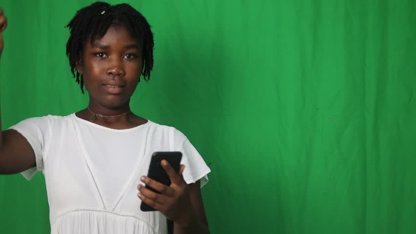 A smiling young woman in a white dress looks excitedly at her phone. She gives a thumbs-up, expressing joy and satisfaction at a good message or digital achievement. The green background reinforces th