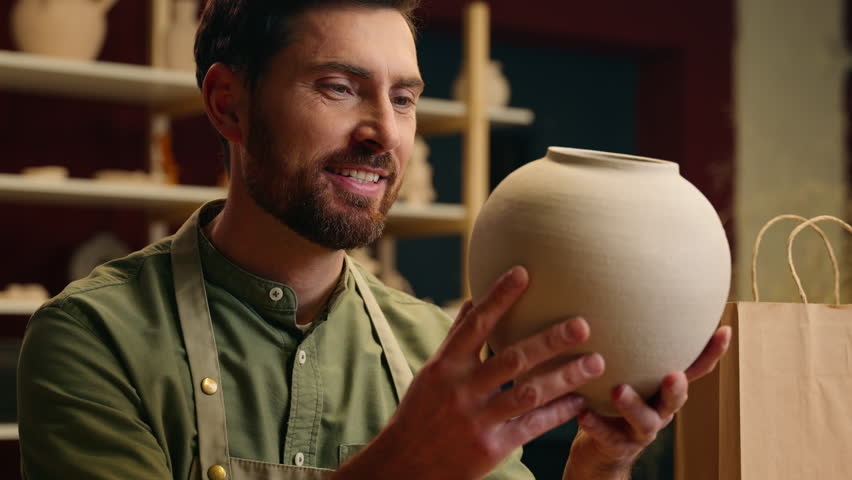 Happy creative smiling Caucasian man guy small business owner businessman crock potter male admiring creation porcelain pottery vase ceramic pot crockery manufacturing packing order in workshop studio