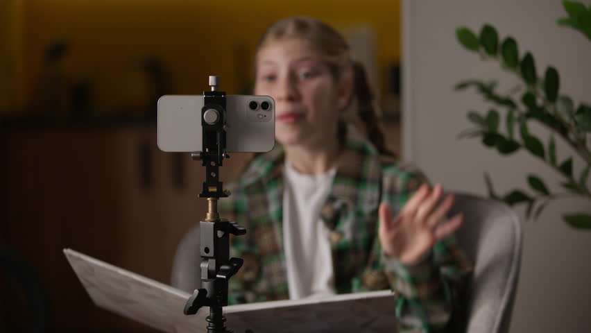 Caucasian teen little girl influencer reading book filming blog smartphone child kid blogger waving hands holding literature read recording audio story podcast using mobile phone camera tripod at home