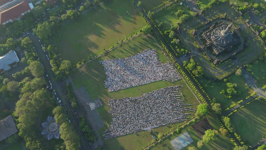 Eid al-Adha Prayer Bali.
Smooth aerial view sweeps over a vast green field, capturing a large Muslim gathering in two rectangular formations for Eid al-Adha prayer next to a temple in Bali.