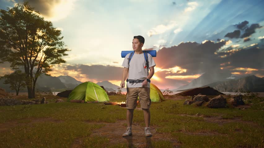 Full Body Of Asian Male Hiker With Mountaineering Backpack Smiling And Pointing To Side while Tent Camp Lakeside