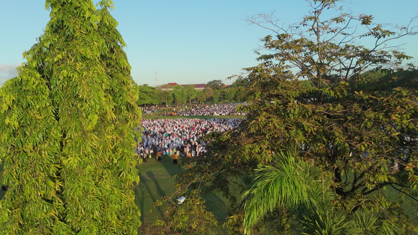 Eid al-Adha Preparation Bali.
Smooth aerial view captures a massive gathering of Muslim people, predominantly in white, preparing for Eid al-Adha prayer on a vast green field in Bali.