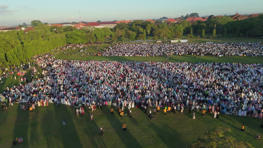 Eid al-Adha Preparation Bali.
Smooth aerial view captures a massive gathering of Muslim people, predominantly in white, preparing for Eid al-Adha prayer on a vast green field in Bali.