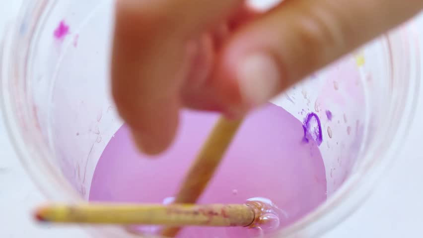 Cleaning paint brush in water in transparent cup. A child washes a paint brush in glass of water after painting. A view from above.