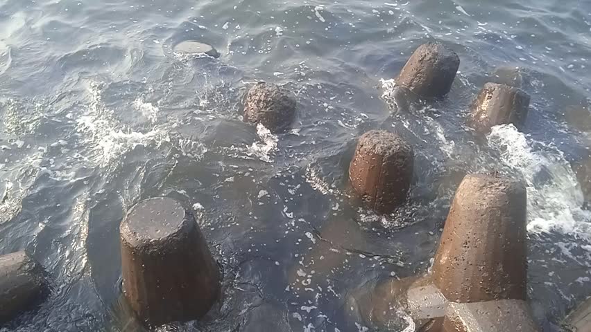 Wave breakers (tetrapods) made of concrete placed along the seashore. The sea waves are crashing against and flowing between the structures, creating splashes and white foam.