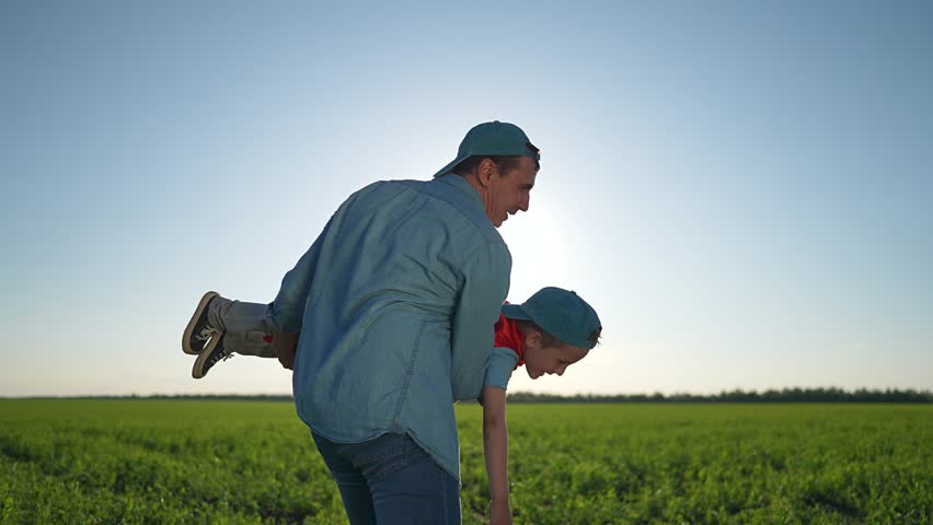 Father holds boy playing superhero bright field. Boy wears cape laughs like cheerful boy. Child stretches arms superhero flying. Green field shines boy joy. Father supports superhero joy, outdoor fun.
