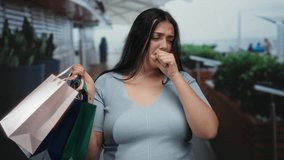Woman coughing outdoors in a city while carrying shopping bags, looking distressed, wearing a light blue top, long black hair, plus size, curvy, standing on a street. - Powered by Shutterstock - Get 15% off with code: PIKWIZARD15