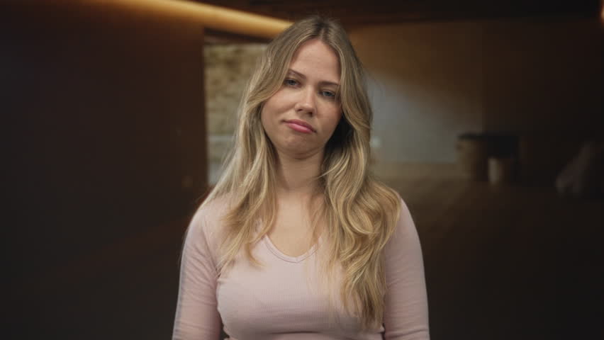 Blonde woman wearing pink top wipes tears at her eyes beside wooden table under soft warm light in room; sadness sorrow vulnerability.