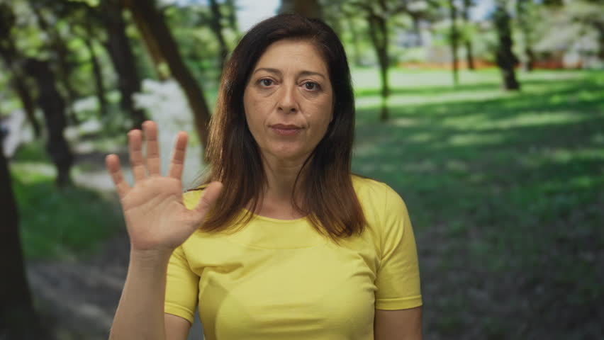 Middle aged hispanic woman wearing a yellow tshirt stands among green trees under sunlight and waves her hand in forest clearing; friendly greeting.