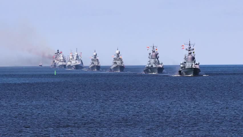 Aerial view of a powerful naval fleet sailing in tight formation across calm blue waters under a clear sky. Modern warships display national flags and emit smoke trails and symbolizing strength.