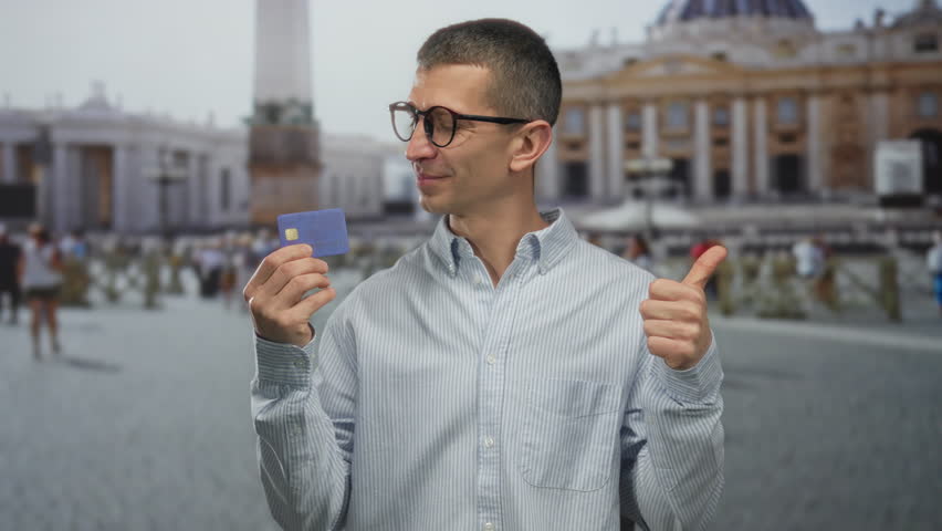 Man holding credit card and showing thumbs up at st. peter's square in vatican city, smiling confidently against a historic backdrop, symbolizing travel and financial stability.
