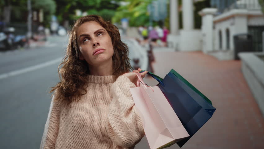 Woman with curly hair making a frustrated gesture with her hand shaped like a gun to her head, holding colorful shopping bags on a city street wearing a casual sweater outdoors.
