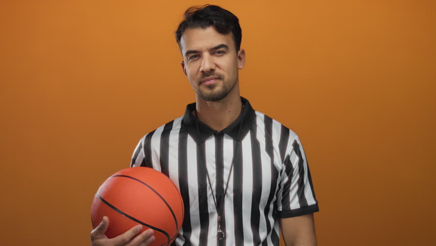 Young hispanic man wearing referee uniform holding basketball against orange background embodying athletic referee persona in isolated vibrant setting.