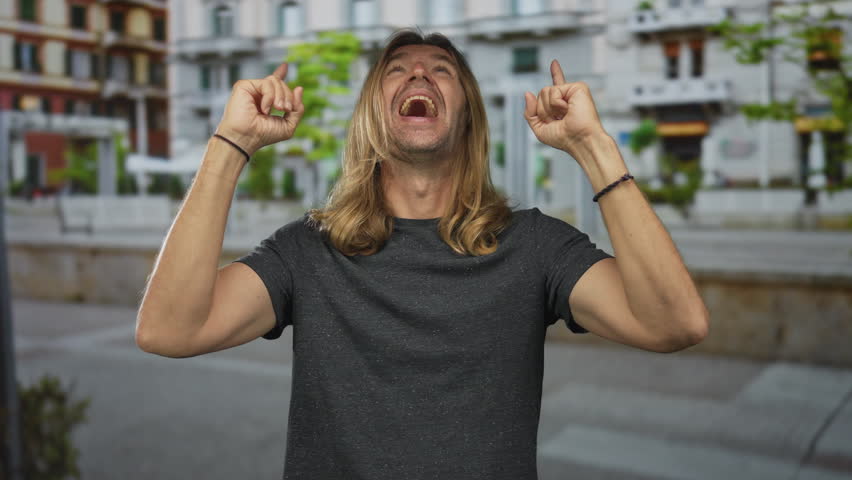 Man with long blond hair pointing both index fingers upward on urban street while smiling broadly in daylight; optimism.
