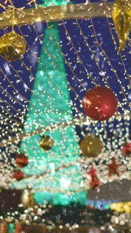 Vertical shot. Christmas european city illuminated with holiday lights. Winter holiday lights illuminate festive decorations in vibrant market in December. First snow falling