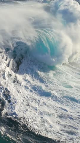 Massive ocean waves crash against rocky shore, creating impressive splashes and mist near coastal cliffs. Aerial slow motion shot of big epic waves surf crash. Vertical shot