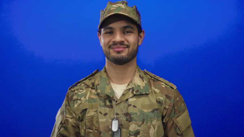 Man wearing camouflage uniform and dogtags points finger to head in studio; confidence reflection focus purpose.