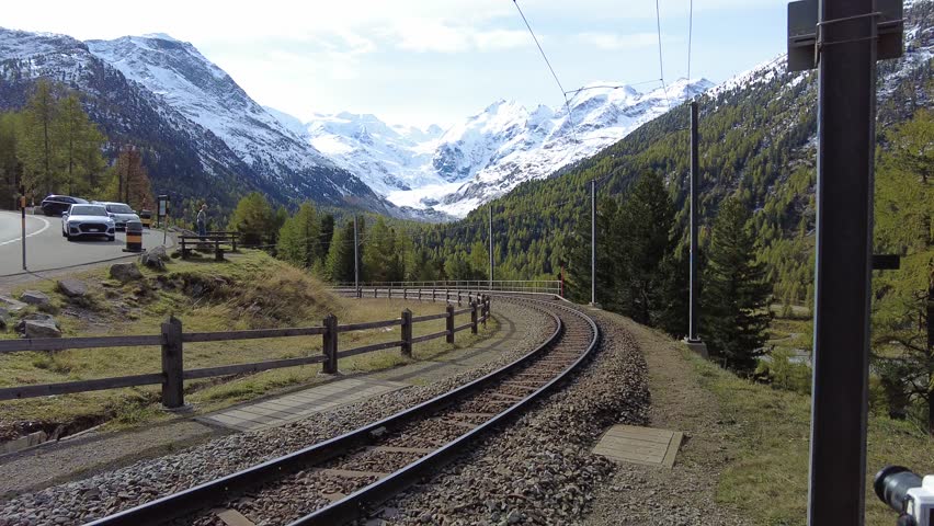 A train of Rhaetian Railway dashing thru Montebello Curve on a beautiful autumn day, with Morteratsch Glacier below Piz Bernina and snowy mountains in background in Pontresina, Graubünden, Switzerland