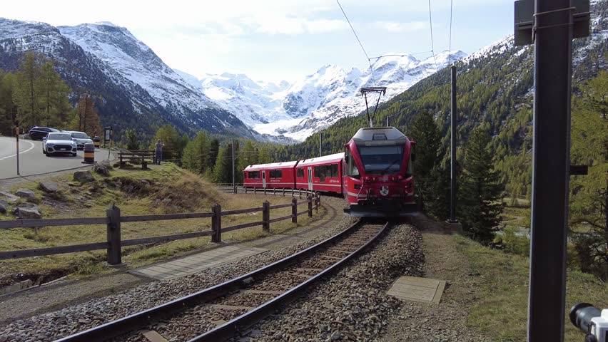 A train of Rhaetian Railway dashing thru Montebello Curve on a beautiful autumn day, with Morteratsch Glacier below Piz Bernina and snowy mountains in background in Pontresina, Graubünden, Switzerland